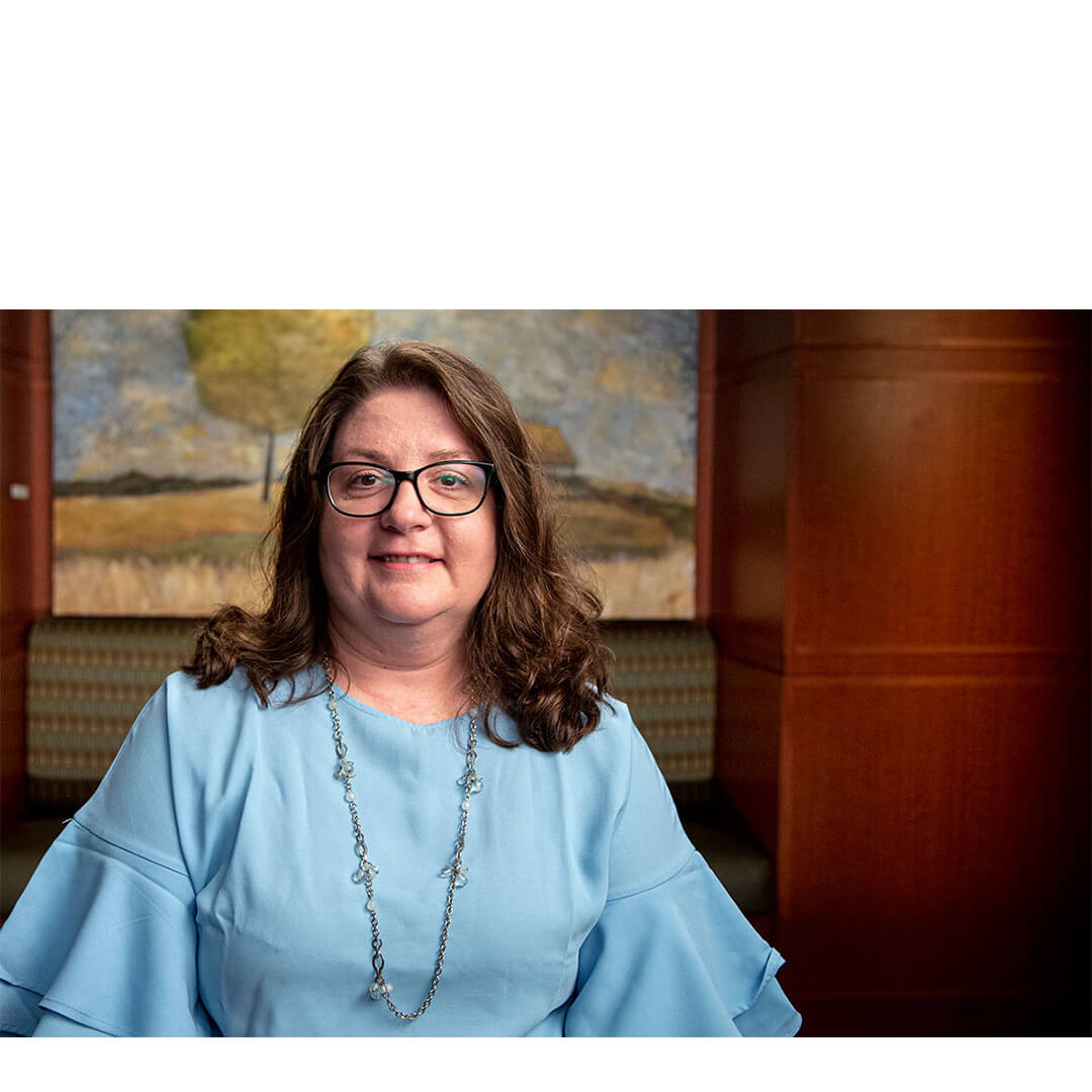 Colleen Walker, a smiling breast cancer survivor wearing glasses and a blue blouse, sits in a wood-toned counseling room, radiating calm confidence