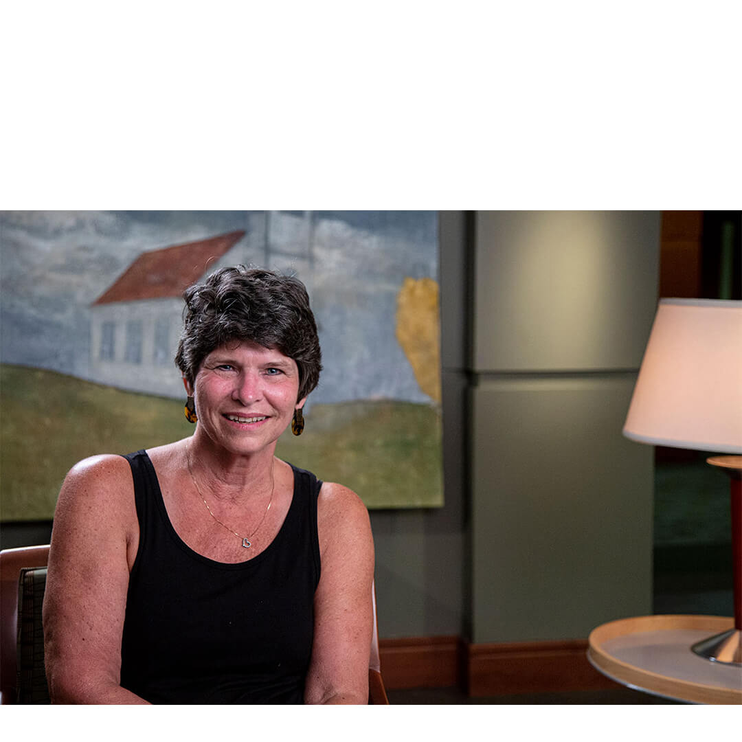 Janice Cheshire, a smiling breast cancer survivor, sits in a softly lit counseling room with a pastoral painting behind her