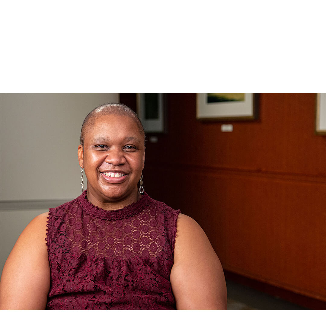 Lomay Richmond, a smiling breast cancer survivor wearing a sleeveless dress and earrings, sits in a warm, wood‑paneled clinic room